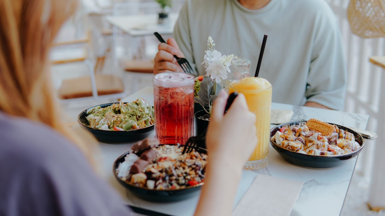 about-01 Two people enjoying a casual meal with various lunch bowls and refreshing drinks at a light-filled café.