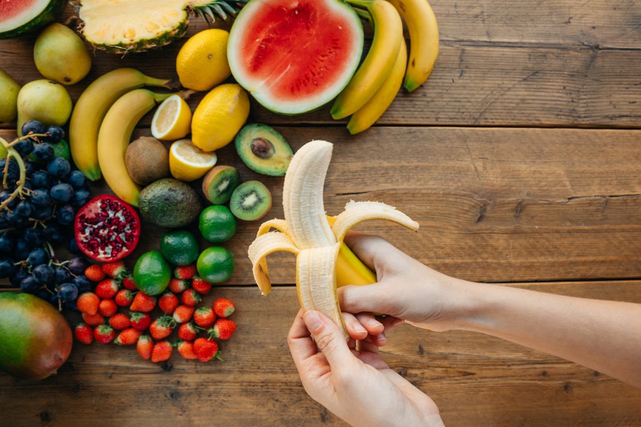 about-02 A person peels a banana surrounded by a variety of fresh fruits on a wooden table.
