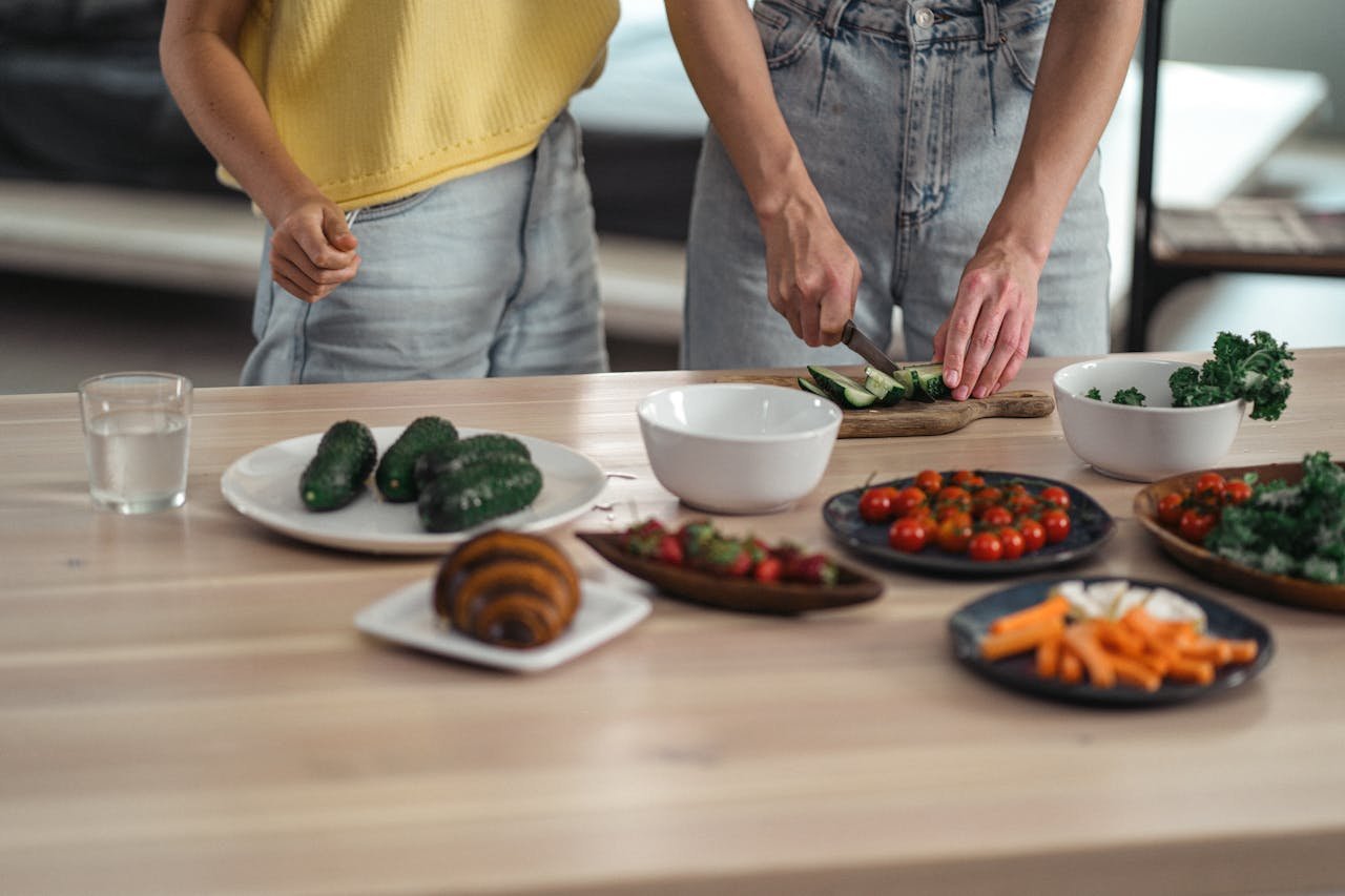 hero-img-02 Two people preparing a healthy vegetarian meal with fresh vegetables and snacks on a wooden table.